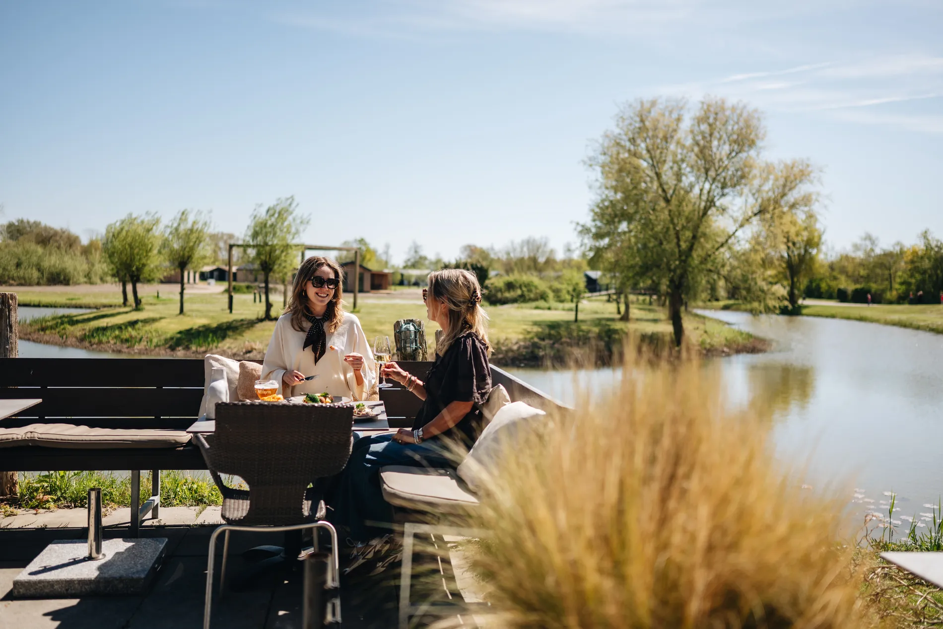 Twee vriendinnen met zonnebrillen lunchen gezellig aan een houten tafel op het terras aan het water, omringd door het weidse, groene landschap van het resort.